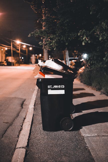 A black plastic wheelie bin with the label 'ST. JOHN'S' positioned on the edge of a paved sidewalk alongside a quiet street at night. The bin's lid is open, revealing various waste items inside, inclu